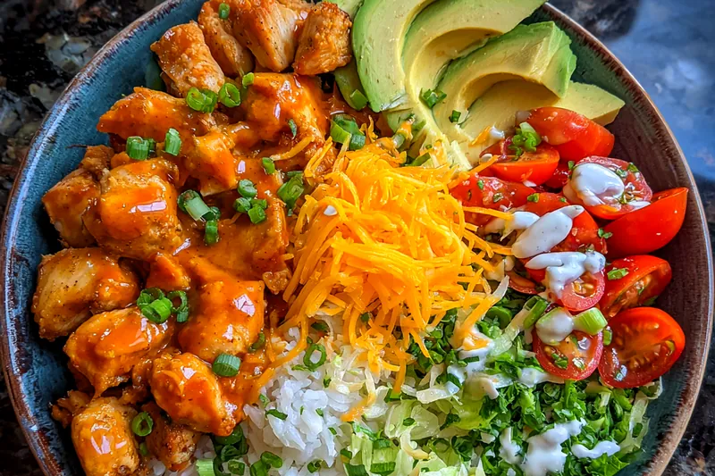 The cooking process showcasing chicken being sautéed in a skillet, surrounded by fresh ingredients for Spicy Buffalo Chicken Bowls.